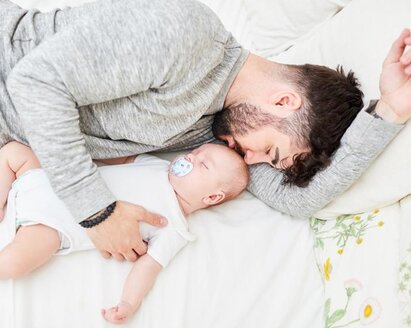 Vater und Baby schlafen im Bett nebeneinander, der Vater hält das Baby.
